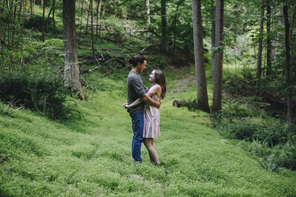 A couple stands facing one another in the lush green forest of the PA woods during an engagement session