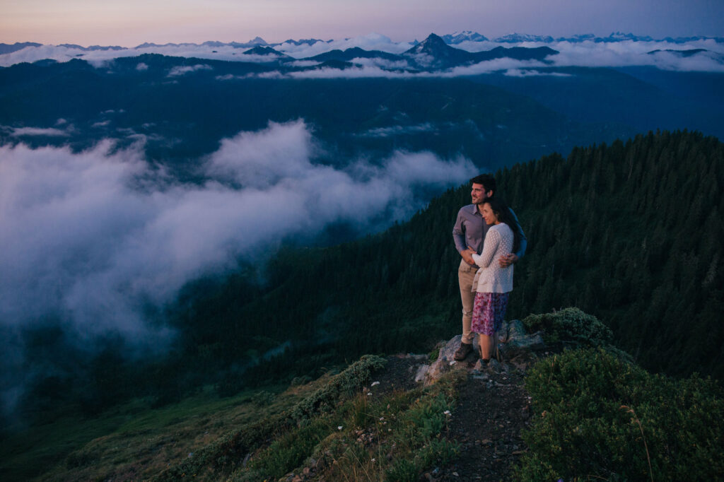 A couple with their arms around one another. The are standing on a summit above the clouds during blue hour. She is wearing a purple floral dress and a white sweater, and he i wearing a blue shirt and tan pants