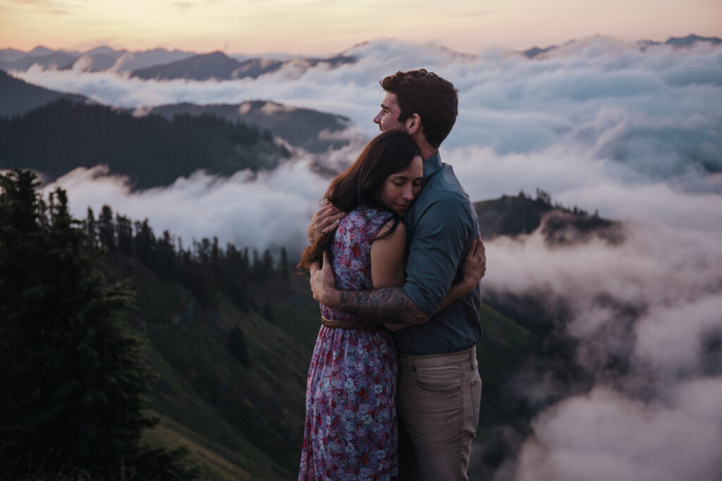 A couple embraces. The woman's eyes are closed with emotion. The are standing on a summit above the clouds before sunrise