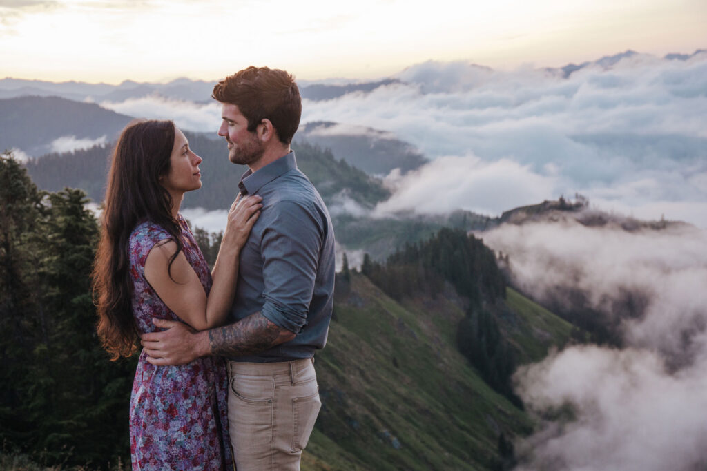 A couple embraces. The husband are gazing into one another's eyes The are standing on a summit above the clouds before sunrise. She is wearing a purple floral dress, and he is wearing a blue long sleeved shirt with tan pants