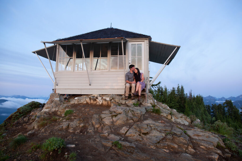 A couple sitting on the stairs of Evergreen Mountain Lookout. Her legs are crossed, and he is kissing her forehead. 