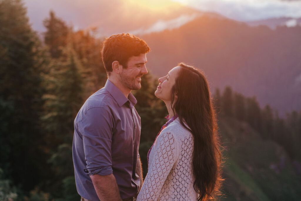 A couple embraces. They are both smiling.  The are standing on a summit above the clouds as the sunrises. The sun is backlighting the scene