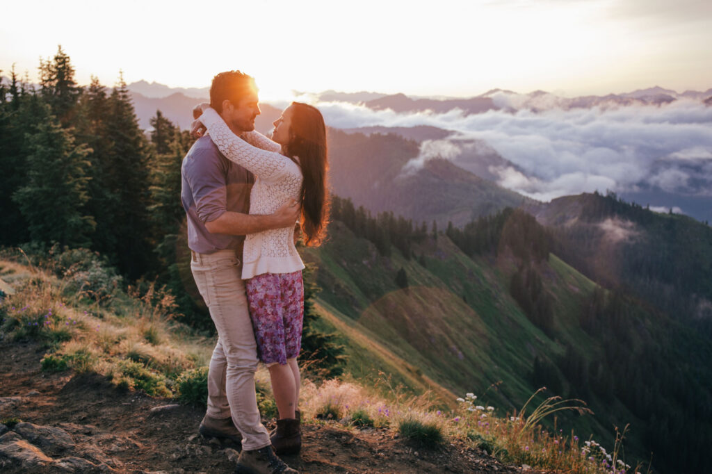 A couple embraces. The woman's arms around the mans neck, and they are smiling.  The are standing on a summit above the clouds as the sunrises. The sun is backlighting the scene