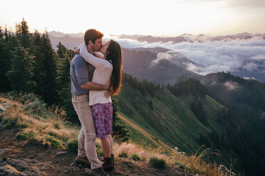 A husband and wife embrace and share a kisswhile standing in front of trees on a summit outside of Seattle Washington. She is wearing a purple floral dress and a white sweater, and he is wearing a blue long sleeved shirt with tan pants.They are standing on the top of the summit above the clouds as the sun rises. 