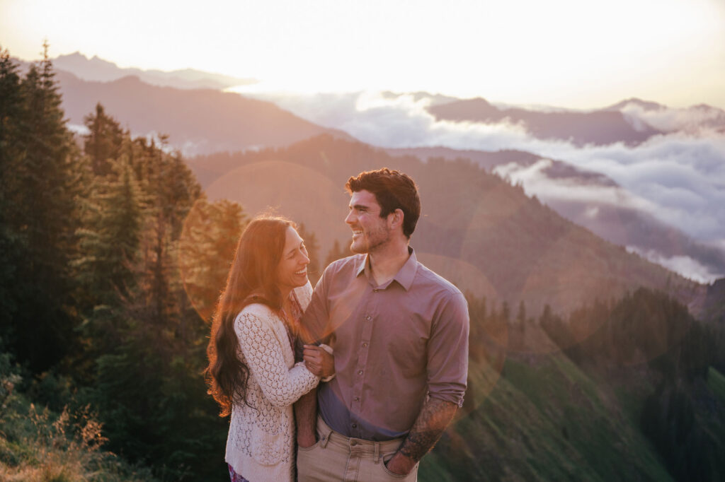 A husband and wife stand next to one another with their arms entwined. They are standing at the top of a summit outside of Seattle Washington. The are both smiling. The sun is rising, and backlighting the photos. There is a sunflare around the couple