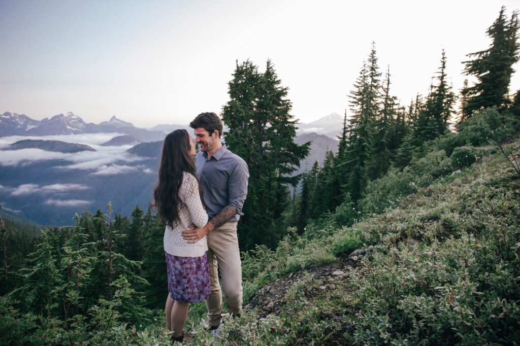 A couple embrace while standing in front of trees on a summit outside of Seattle Washington. She is wearing a purple floral dress and a white sweater, and he is wearing a blue long sleeved shirt with tan pants. They are looking at one another and smiling