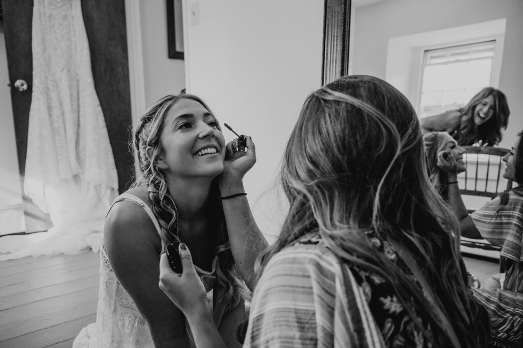 A woman getting her makeup done on her wedding day. The photo is in black and white, and the woman is smiling as her friend puts mascara on her.