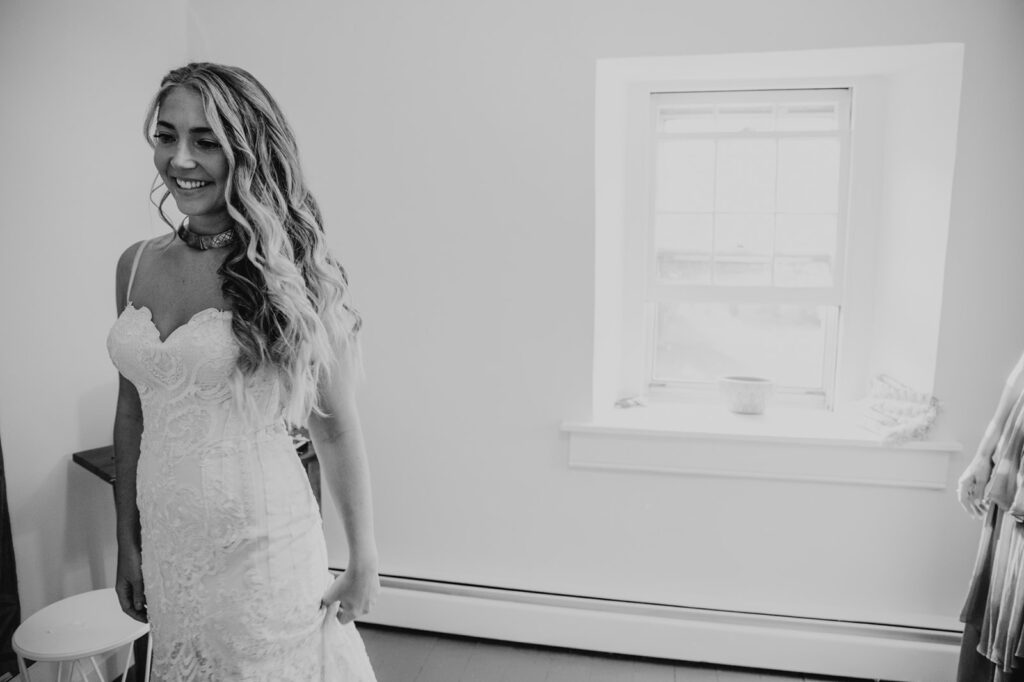 A black and white photo of a bride seeing herself for the first time in the mirror. The bride is wearing a sleeveless dress.