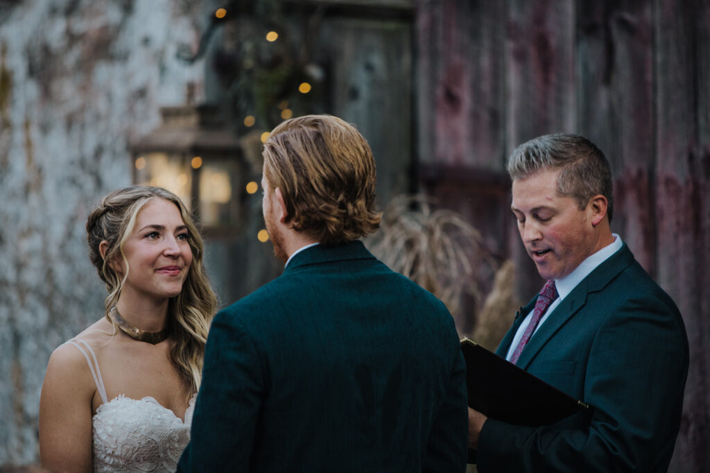 A bride looks at her groom lovingly during their small wedding ceremony. The bride is wearing a sleeveless lace dress, and the groom is wearing a blue jacket.