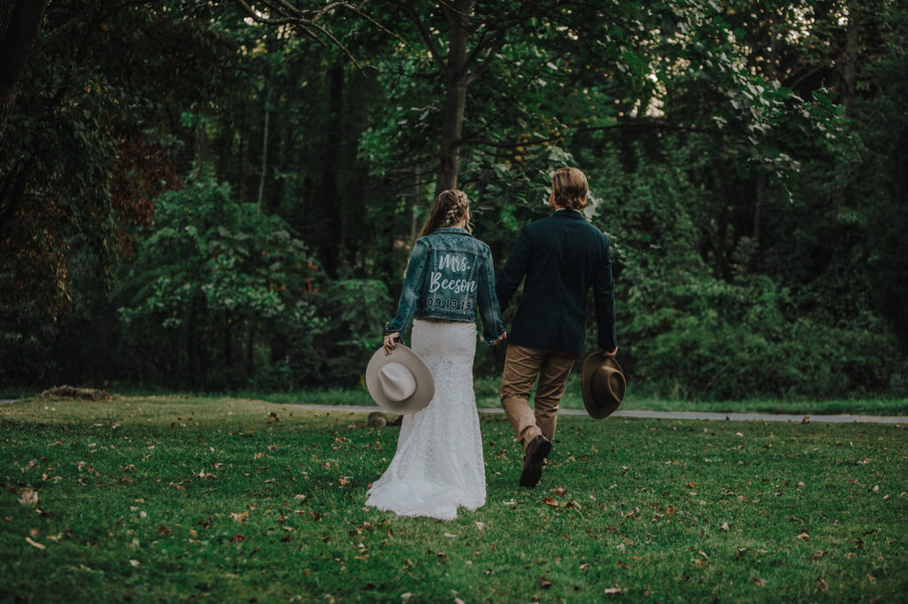 A bride and groom walk hand in hand away from the camera. The bride is wearing a custom denim jacket, and they are both holding cowboy hats