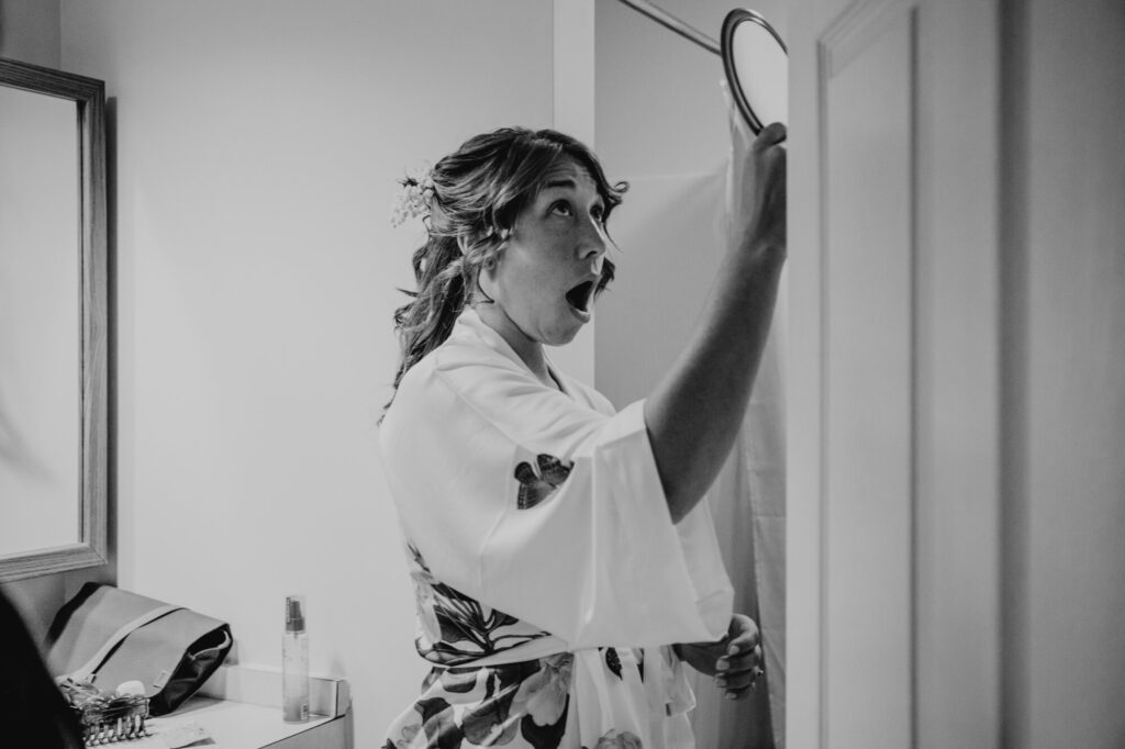 A black and white photo of a bride seeing her hair and makeup in a mirror for the first time. She has her mouth open in shock, like"gosh I look good"