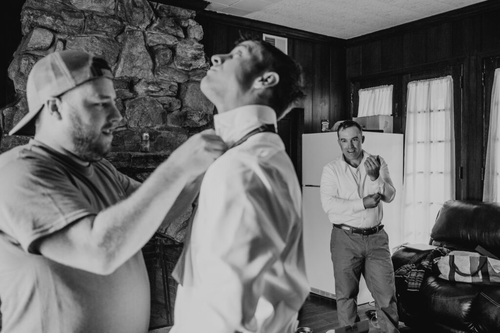 A black and whit photo of three men getting dressed for a wedding. The groom is in the background buttoning his sleeves, while a man in the foreground helps another man with his tie.