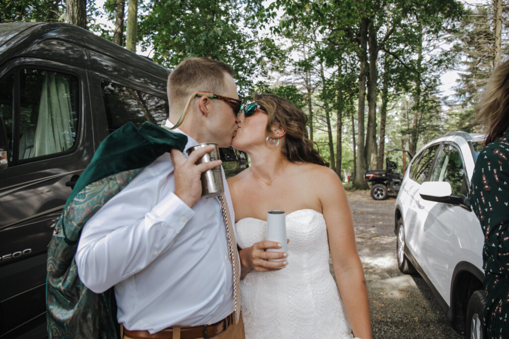 A bride and groom, both wearing sunglasses kiss in a campground parking lot. Both are holding their insulated cups. The bride is wearing a strapless dress, and the groom has his green velvet coat over his shoulder