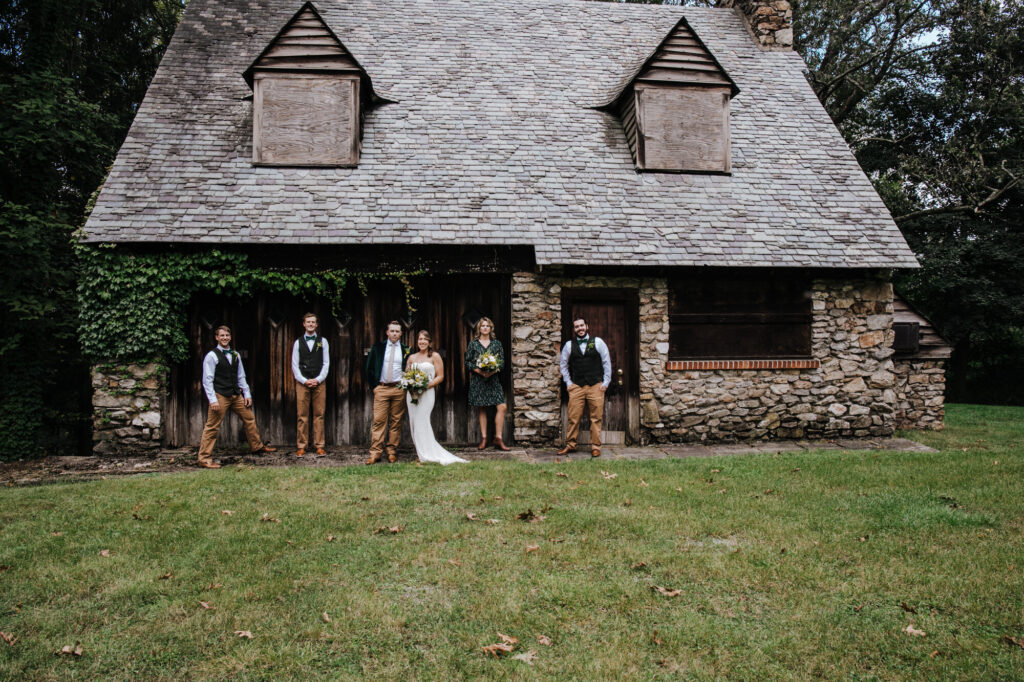 A couple in front of a lodge the woman is wearing a white dress the man is wearing a white dress jacket and tan pants and they are walking together on a lawn and their wedding party is behind them watching them