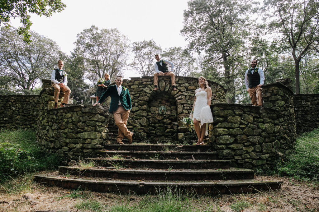 A bride and groom and their wedding party pose for a photo on an old stone wall and steps. The bride is holding up her dress, and the rest of the group is casually posed around her. 