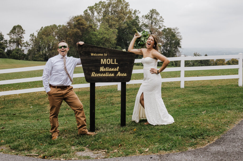 A woman in a white wedding dress is laughing and holding her bouquet in the air and a man in a white shirt and tan pants who is also  wearing sunglasses is smiling and they are both standing next to a wooden sign 