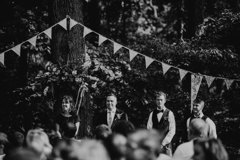 A black and white photo of a groom, the officiant, and two groomsmen, watching the bride walk down the aisle towards them. There is a pennant banner hanging in the background