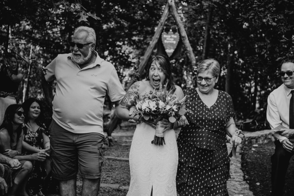 A black and white photo of a bride walking down the aisle with her parents. The bride has a look of excitement on her face.