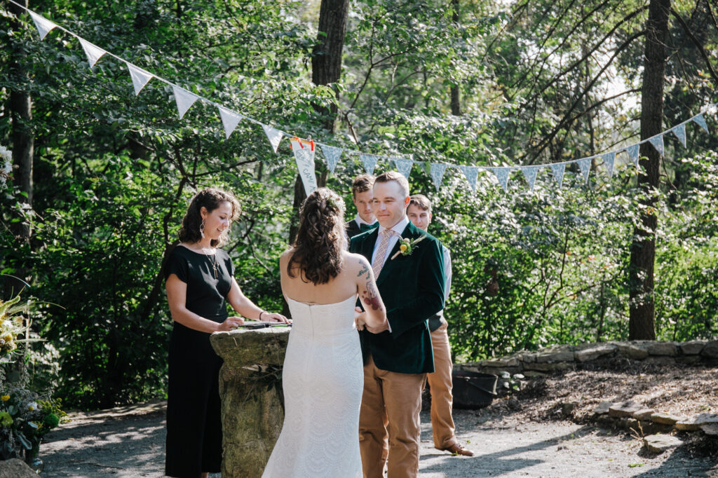 A bride and groom hold hands while facing each other during the vows of their ceremony. The bride is wearing a strapless dress, and the groom is wearing a green velvet jacket. There is a pennant banner hanging in the background.