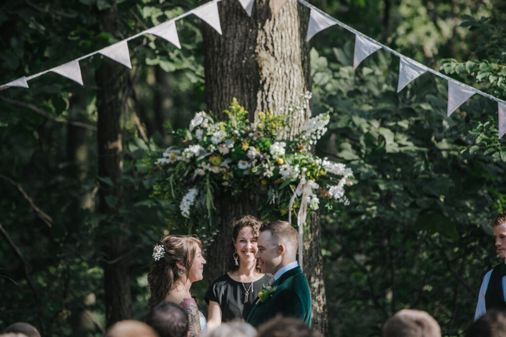 A bride and groom hold hands while facing each other during the vows of their ceremony. The bride is wearing a strapless dress, and the groom is wearing a green velvet jacket. There is a pennant banner hanging in the background, and a large floral arrangement.