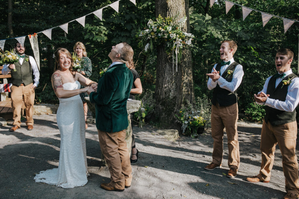 A bride and groom hold hands while facing each other during the vows of their ceremony. The bride is wearing a strapless dress, and the groom is wearing a green velvet jacket. The bride and groom are both laughing. There is a pennant banner hanging in the background.