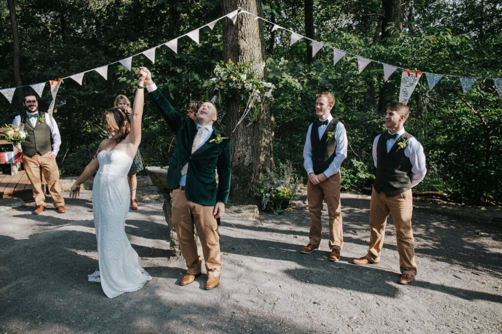 A bride and groom throw their hands in the air in celebration of officially being married. Their bridal party is standing behind them smiling.