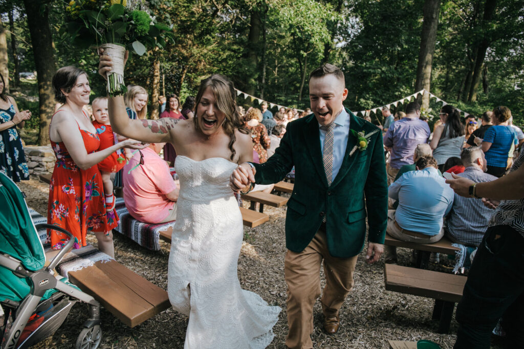 A bride and groom exit their ceremony celebrating officially getting married. Their guests are standing and cheering. The bride is wearing a strapless dress and the groom is wearing a green velvet jacket.