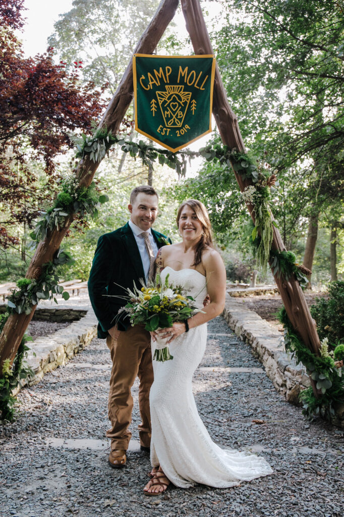 A woman in a white wedding dress is holding a bouquet of flowers and smiling and a man in a blazer and tan pants is smiling and standing next to her and behind them is a wooden arch