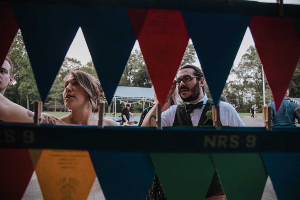 A woman and man look at the seating chart for their seat at a wedding reception. The camera is shooting through pennants, and they are framing the man and woman