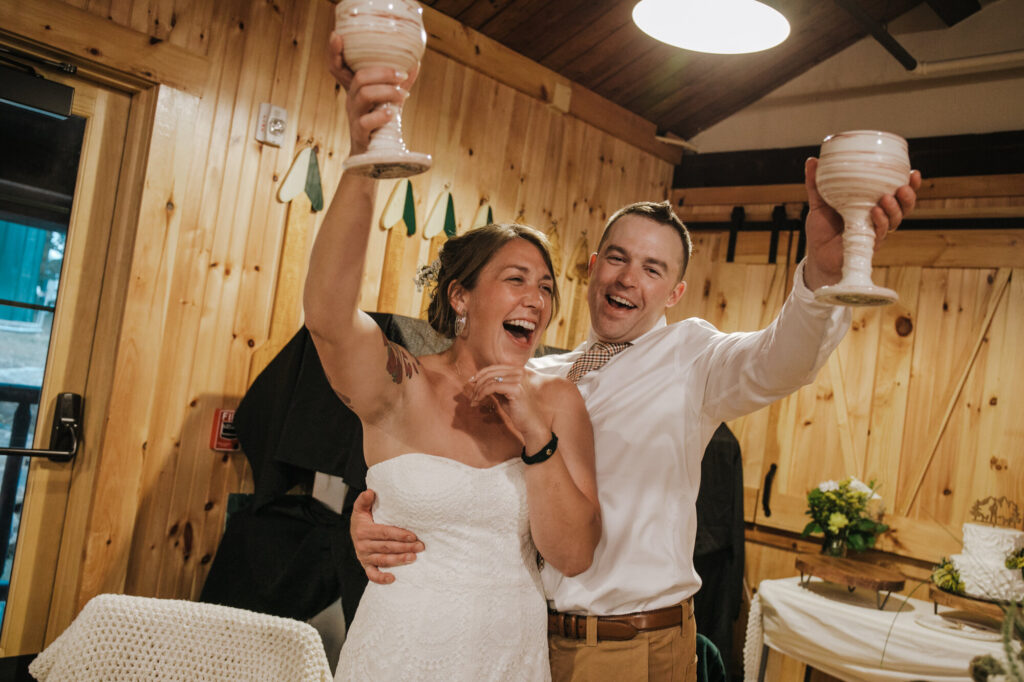 A bride and groom raise their glasses during a toast. The glasses are hand made goblets. The couple is laughing at the toast