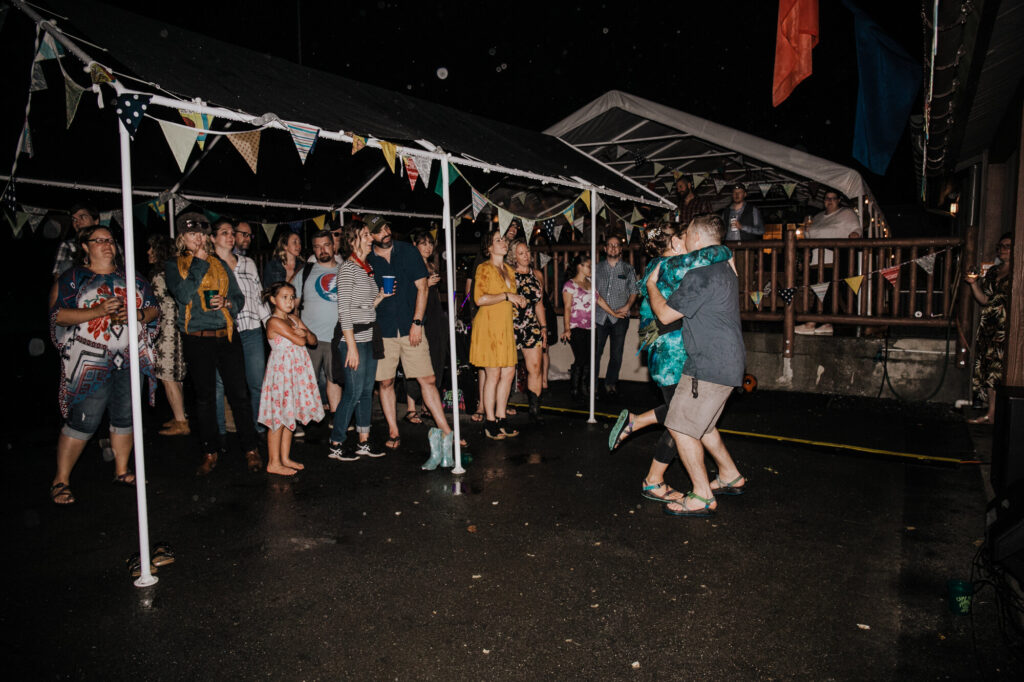 A woman and man dance in the rain, as their friends watch. The couple is in the rain, and their friends are standing under a tent decorated with a colorful flag banner. The woman in wearing a teal tie dye sweatshirt, and the groom is wearing  a black shirt, and brown shorts