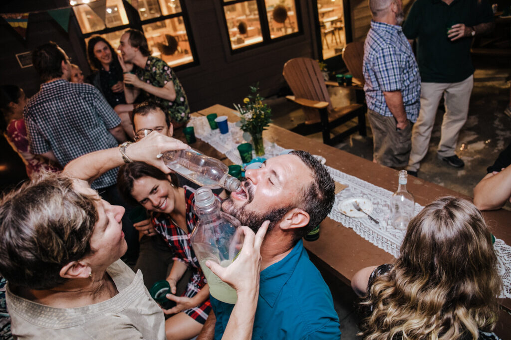 A woman is pouring tequila into a mans mouth while he is seated at a picnic table