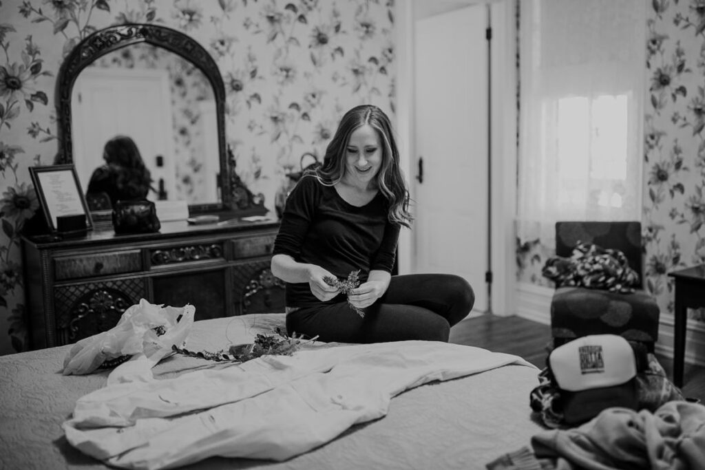 A bride making bouquets and boutonniere's on her wedding day. She is sitting on  the bed along with her jumpsuit that she is going to put on later.