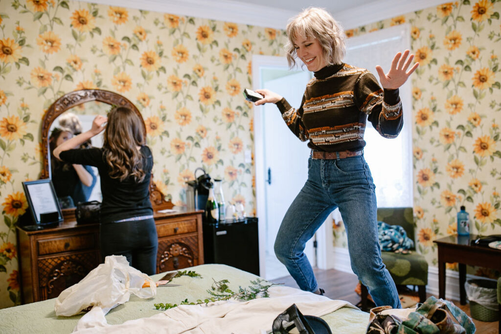 A girl jumping up on the bed in a zany and fun way. The bride is also in the room doing her hair in front of the mirror.