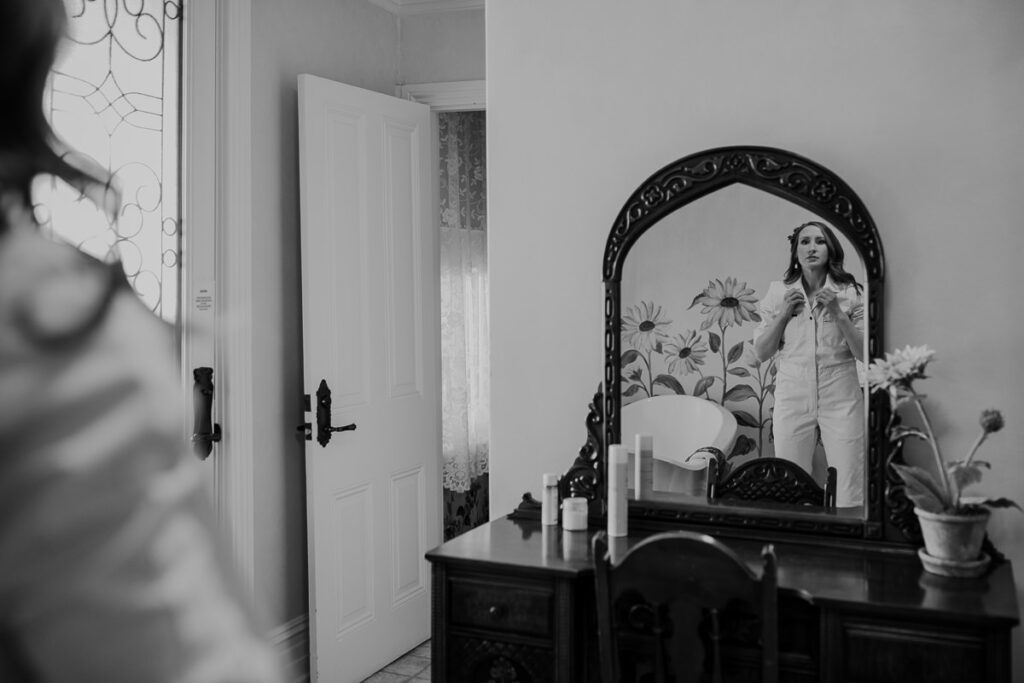 A bride looking at her herself in the mirror while she finished getting dressed. She is wearing a white Lee jumpsuit.