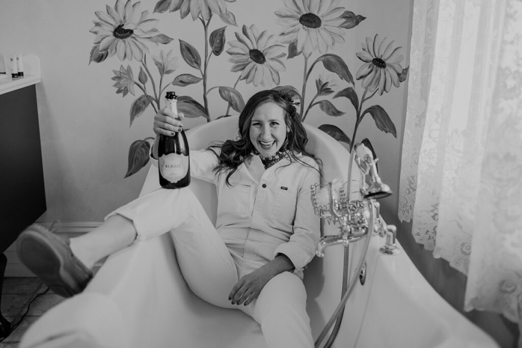 A black and white photo of a bride in a bathtub. She is wearing a Lee jumpsuit, and holding a bottle of champagne. Her leg is hanging out of the bathtub. Their are sunflowers painted on the wall behind her