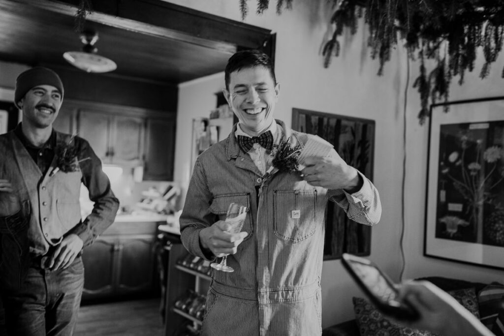 A groom in a pinstripe jumpsuit and a bowtie laughs while in his home. He is also holding a glass of wine,