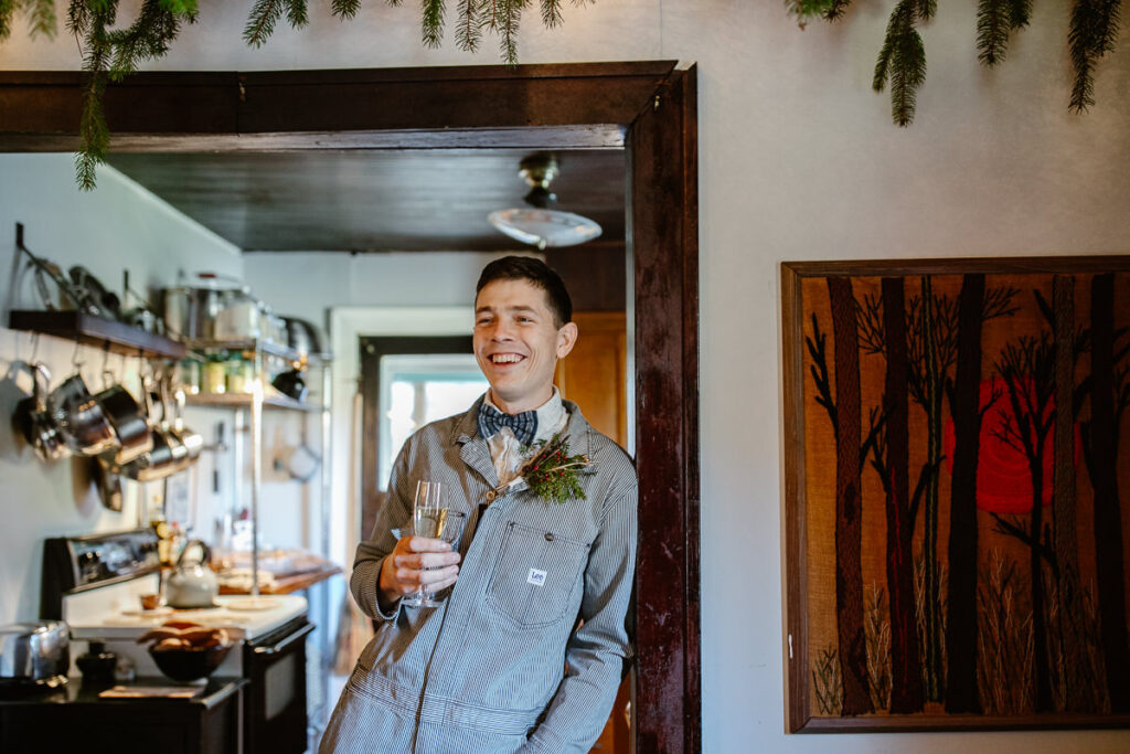 A groom wearing a pin striped Lee jumpsuit leans against the wall holding a glass of wine