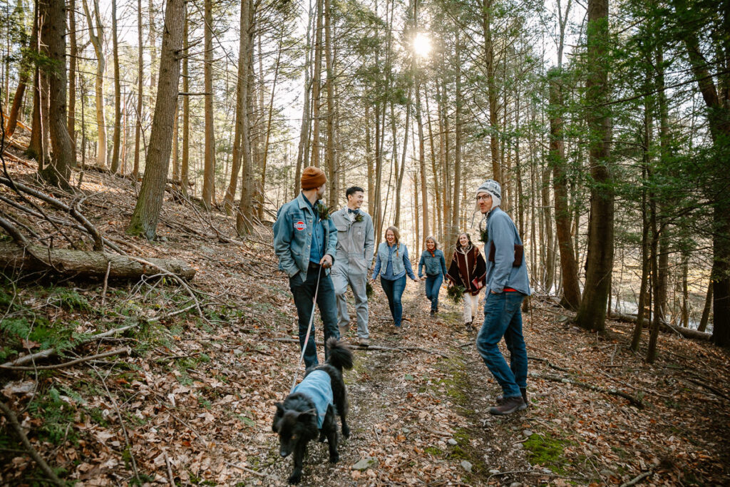 A wedding party dressed in all denim is walking up a hill in the woods  to a ceremony location. A black fuzzy dog is leading the way. The sun is setting behing them and the trees