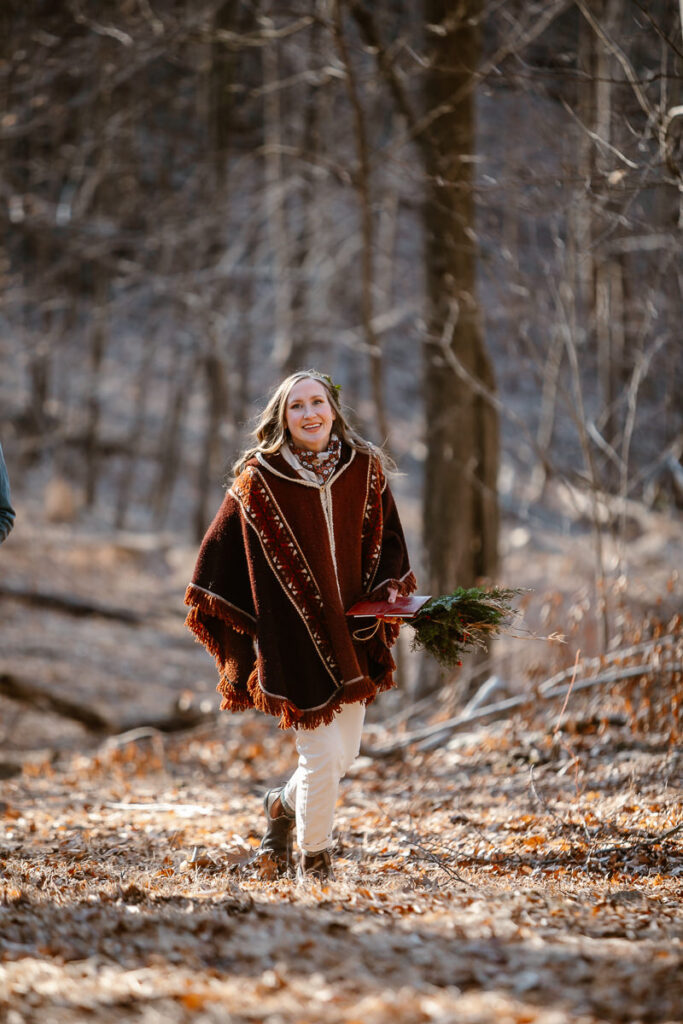 A bride in a poncho and white jumpsuit walks up a hill in the woods.