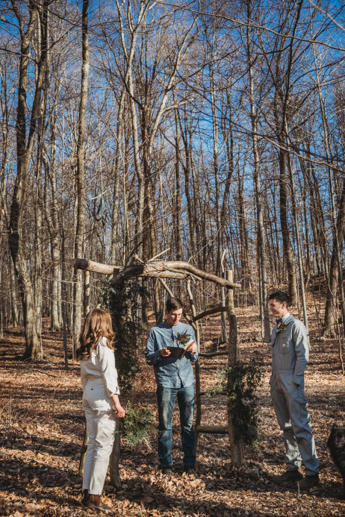 A bride and groom and officiant stand under a hand made arbor in the PA woods. The officiant is wearing all denim, and the bride and groom are wearing Lee denim jumpsuits.