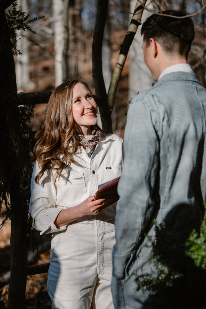 A bride looks up at her groom with a loving smile during their vows. She is wearing a white Lee denim jumpsuit. They are int he woods