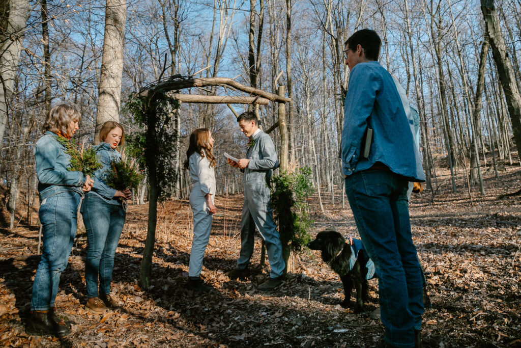 A photo of an elopement in the woods. It is a couple standing under an arbor. Their four closest friends and a dog are all there wearing denim