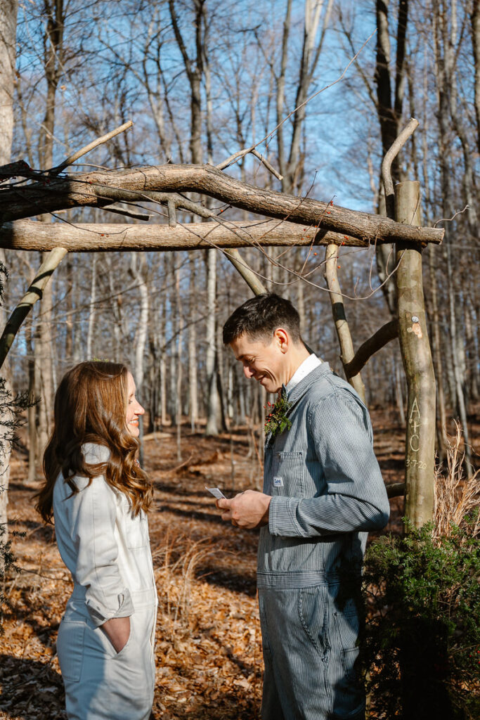 A bride looks at her groom while he reads his personal vows to her on their elopement day. They are both wearing Lee denim jumpsuits. They are standing under an arbor in the woods