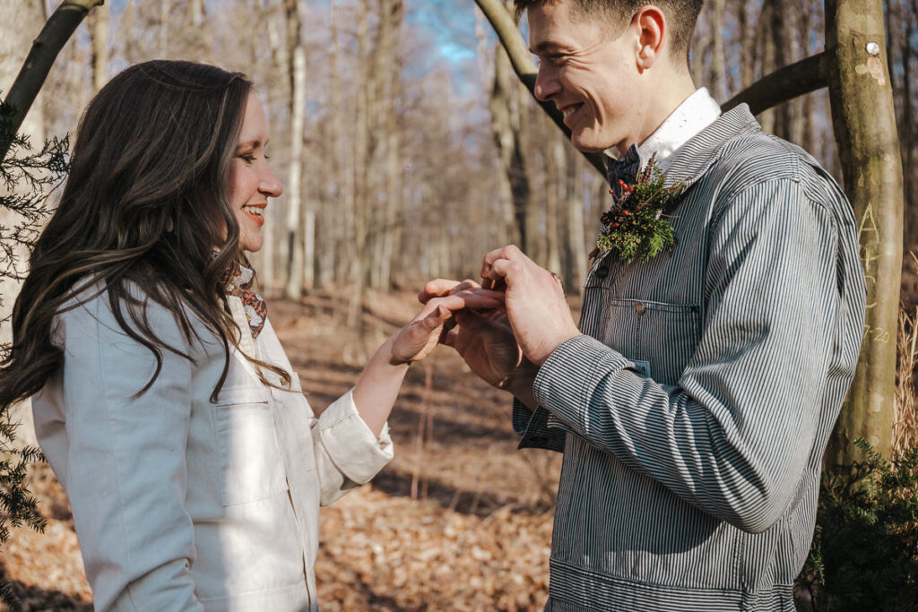 A groom puts a wedding ring on his brides finger during their elopement. They are both wearing denim jumpsuits. 