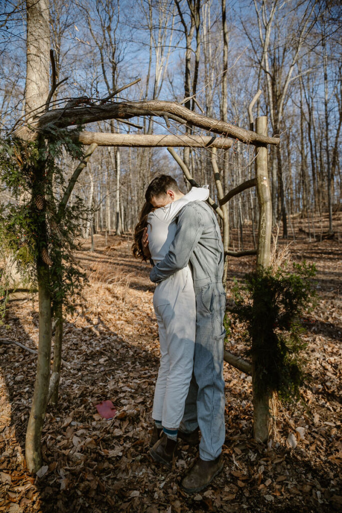 A bride and groom embrace during their elopement. They are standing under an arbor in the woods. They are both wearing denim jumpsuits