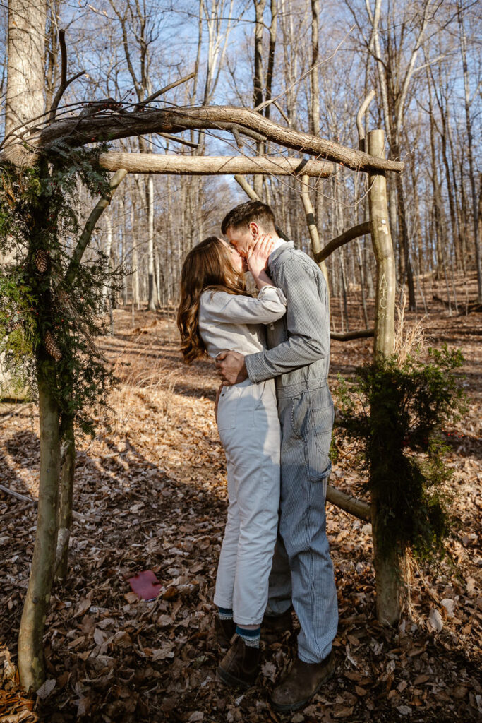 A bride and groom share their first kiss during their elopement. They are standing under an arbor in the woods. They are both wearing denim jumpsuits