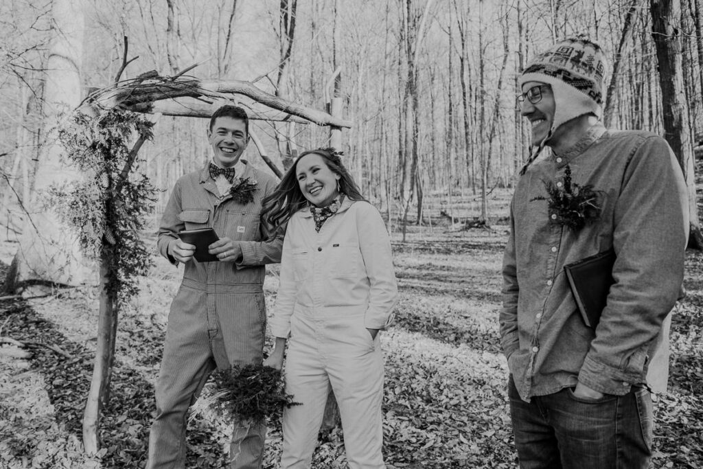 A bride a groom are beaming after their self uniting ceremony. They are both wearing jumpsuits, and standing by an arbor in the woods