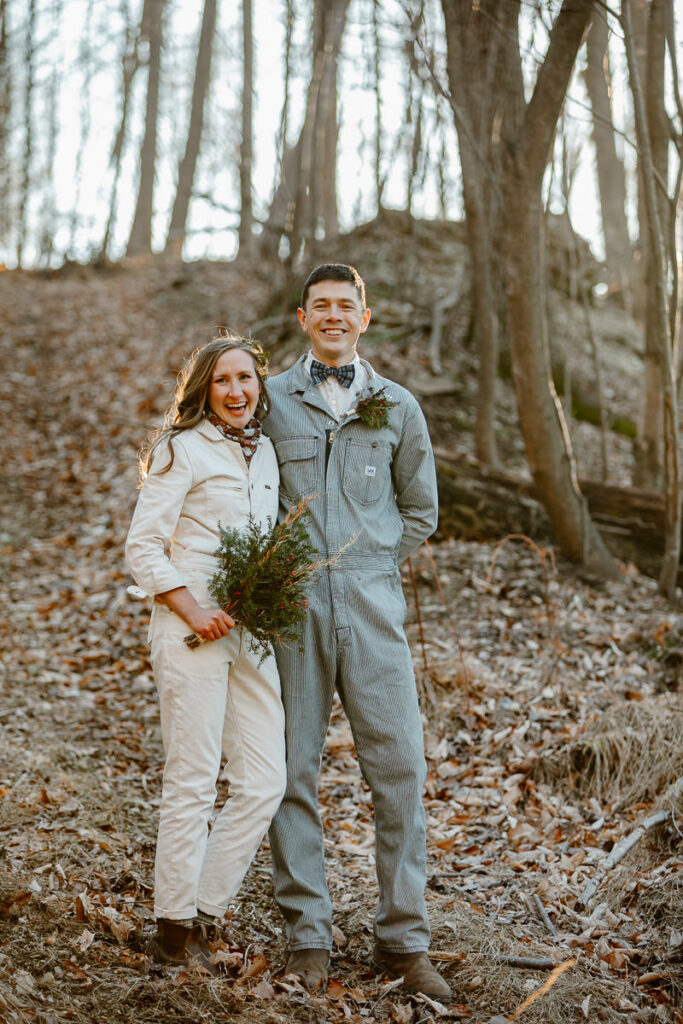 A bride and groom stand smiling at the camera. They are both wearing denim jumpsuits. The photo is backlit from the sun setting