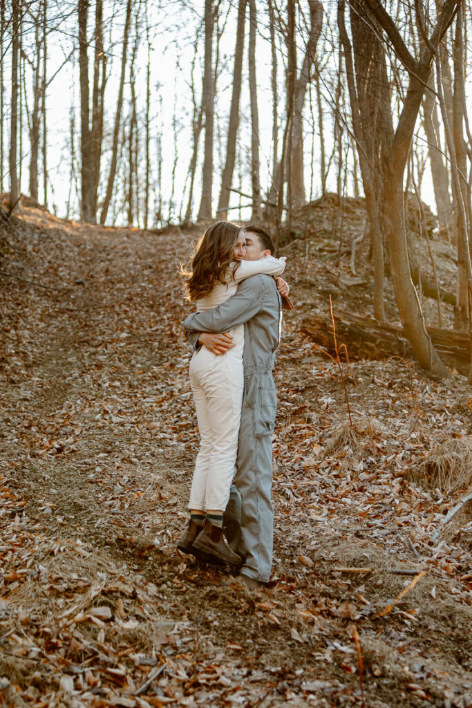 A groom picks up his bride and kisses her cheek. They are both wearing denim jumpsuits. The photo is backlit from the sun setting